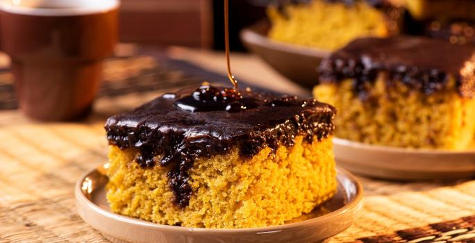 Fotografia em tons de marrom com uma prato pequeno ao centro. Em cima do prato existe uma fatia de um bolo de cenoura de cor alaranjada e com cobertura de chocolate. Ao fundo existe mais fatias desse mesmo bolo.