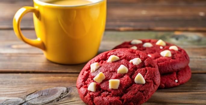 Fotografia de três cookies de red velvet com gotas de chocolate branco juntos, sobre uma mesa de madeira escura, ao lado de uma xícara grande amarela.