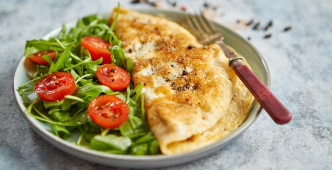 Foto da Receita de Omelete Baveuse. Observa-se um prato com a omelete dobrada no meio, servida com salada de rúcula e tomatinhos cerejas cortados ao meio.