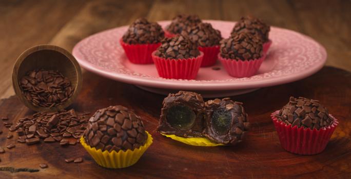 Fotografia em tons de amarelo e vermelho em uma bancada de madeira escura, um prato redondo rosa com brigadeiros em forminhas vermelhas em cima dele. Ao lado, brigadeiros em forminhas amarelas recheados com uva.