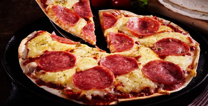 Fotografia em tons de marrom com uma pizza feita com massa de tortilha ao centro. A pizza é feita de queijo, salame e orégano.