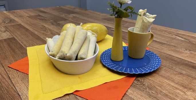 Foto da receita de Sacolé de Maracujá, servido em saquinhos, em tom amarelo, em uma travessa branca, sobre uma bancada de madeira decora com um tecido amarelo e outro laranja, ao lado há um prato azul redondo com um vaso e uma caneca amarelos