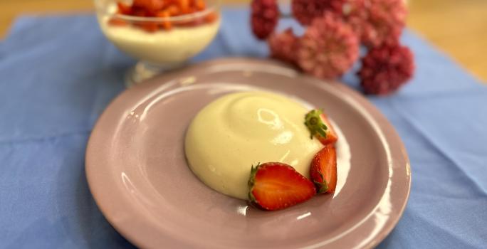 Fotografia em tons de rosa e azul com um prato rosa ao centro. Em cima do prato existe uma receita de Panna Cotta com morangos fatiados por cima