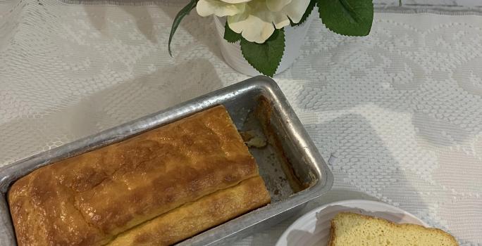 Fotografia vista de cima de um pão dentro de uma forma pequena retangular de alumínio ao lado de um prato pequeno raso com uma fatia do pão.