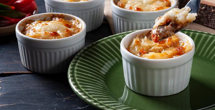 Fotografia em tons de marrom com vários ramekins ao centro. Dentro de cada recipiente existe uma receita de escondidinho de carne seca sem lactose. Um prato de mandiocas gratinadas com carne seca.