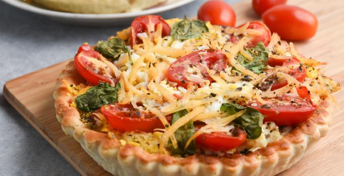 Imagem de uma quiche sobre uma tábua de madeira. A massa da quiche é dourada e crocante e ela está recheada com tomates-cereja, folhas de manjericão e lascas de queijo parmesão.