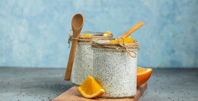 Fotografia em tons de azul com dois copos de vidro ao centro. Dentro de cada pote existe uma receita de Overnight Chia feita com laranja, chia e coco. Ao fundo existe uma laranja cortada ao meio