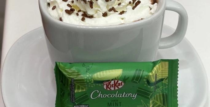 Fotografia de uma xícara branca com um hot chocolate com chantilly e granulado por cima. Na frente da xícara tem um kitkat de menta, e está sobre um prato pequeno de vidro raso branco.