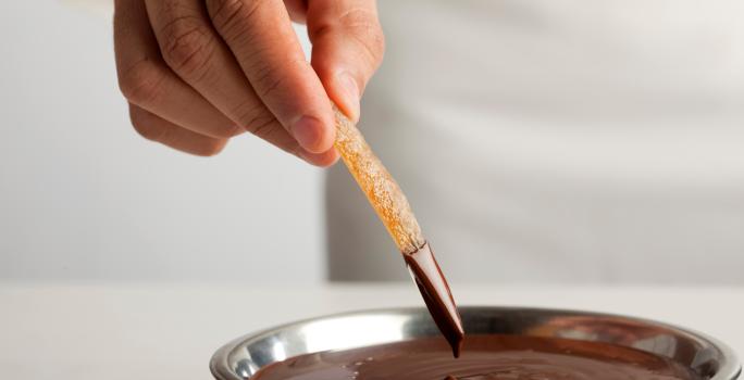 Fotografia de fruta seca afundando no chocolate amargo, que está em um recipiente de alumínio fundo. O qual está sobre uma mesa branca.