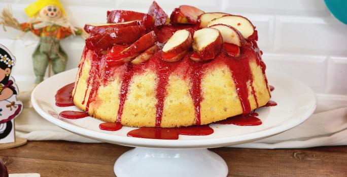 Foto da receita de bolo de maçã do amor, servido em um prato alto branco, com calda vermelha de maçã do amor escorrendo pelas laterais, com pedaços de maçã decorando o topo e itens de festa junina sobre a bancada ao lado do bolo