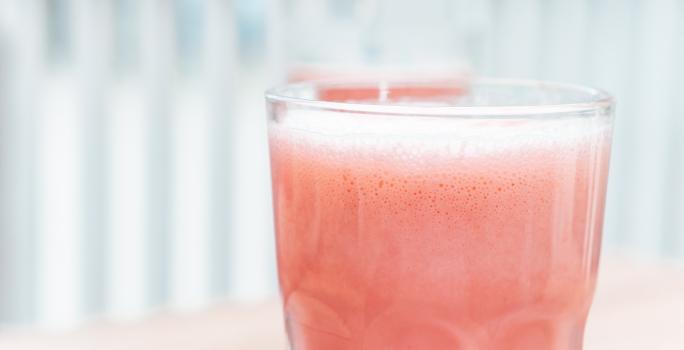 Fotografia em tons claros de uma bebida com um pouco de espuma de melancia com leite MOÇA e neston, que está em um copo de vidro.