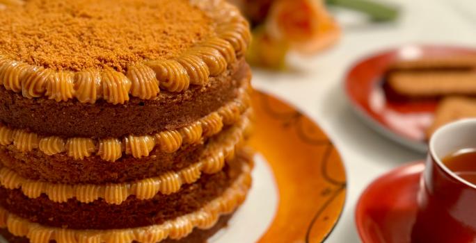 Fotografia em tons de caramelo com um prato de cor marrom ao centro. Em cima do prato existe um bolo com várias camadas de doce de leite e coberto com biscoito triturado