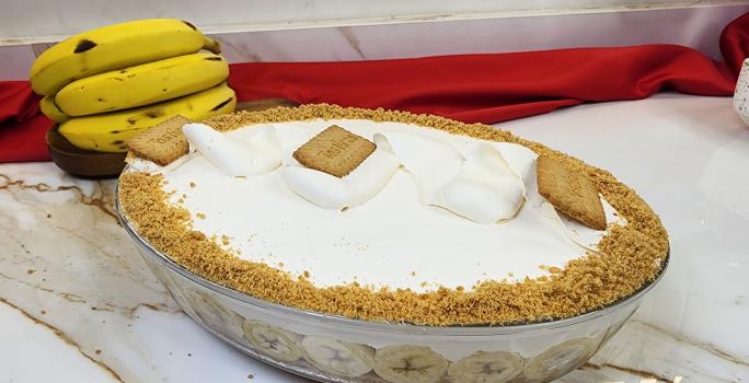 Foto da receita de Torta Banoffee de Travessa, servida em uma travessa oval de vidro, decorada com chantilly batido e biscoitos tostines triturados, tudo em uma bancada de mármore com algumas bananas ao fundo e um tecido vermelho