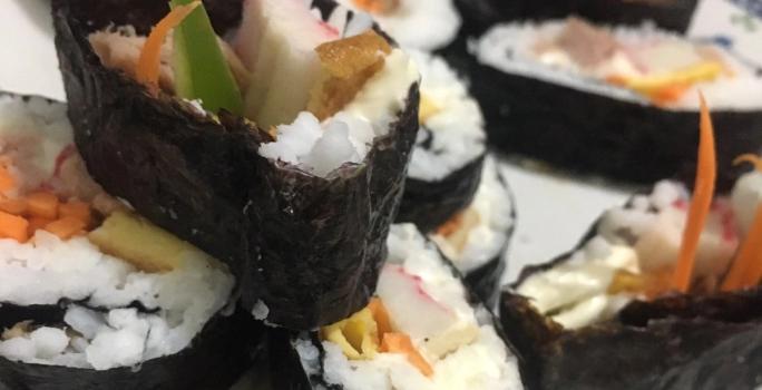 Fotografia de fatias grossas de gimbap brasileiro com arroz, cenoura, kani e pepino sobre um prato branco.