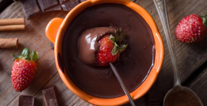 Fotografia mostra um morango mergulhado em uma panela laranja com chocolate derretido.