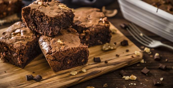 Fotografia de quatro pedaços de brownie feito de farinha de soja, castanha e chocolate. Os pedaços estão sobre uma tábua de madeira perto de um garfo.