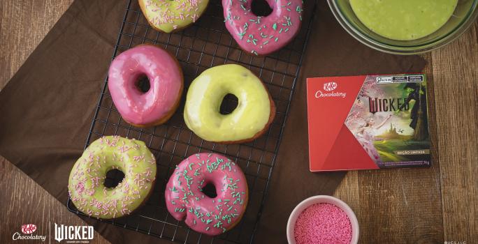 Foto da receita de Donuts Wicked, vistos de cima, servidos em uma grade de ferro preta. São seis no total, decorados com cobertura rosa e verde e alguns com granulados coloridos. Ao lado direito há uma caixa de KitKat Wicked.