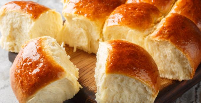 Fotografia de várias unidades pãos fofinhos de leite ninho dourados por cima. Os pães estão sobre uma tábua de madeira.