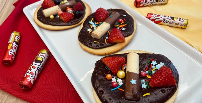 Fotografia em tons de vermelho e amarelo com um recipiente branco ao centro. Em cima do recipiente existe 3 mini pizzas doces cobertas com ganache de chocolate meio amargo, morangos e BATON Duo. Ao lado existe algumas embalagens de BATON Duo