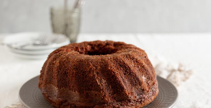 Fotografia de um bolo de chocolate feito em forma com furo central sobre um prato raso de vidro cinza. Ao lado tem granulado de chocolate em um prato pequeno raso. No fundo tem pratos brancos e garfo, e o bolo está sobre um pano branco.