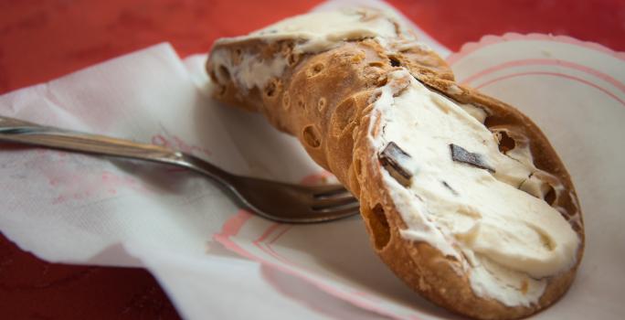 Fotografia de um cannoli de chocolate com creme branco sobre um papel, e ao lado de um garfo.