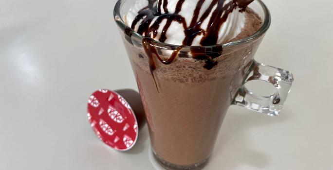 Fotografia em tons de branco com uma caneca de vidro ao centro. Dentro da caneca existe uma receita de frappuccino de cor chocolate coberto com chantilly e calda de chocolate. Ao lado existe uma cápsula de NESTLÉ Dolce Gusto KitKat