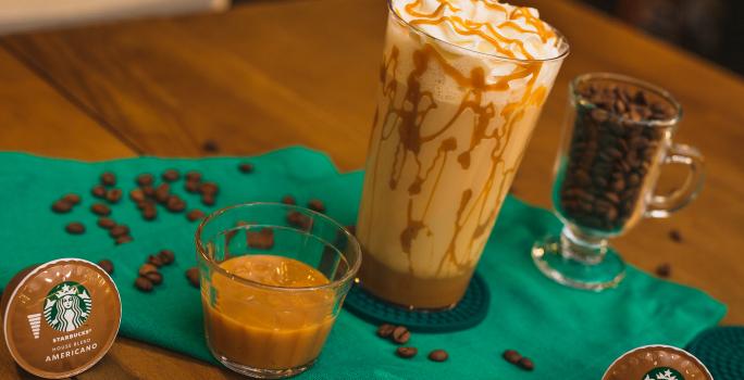 Fotografia em tons de verde de uma mesa de madeira com um paninho verde e ao centro um copo decorado com doce de leite, com uma bebida de leite e decorado com chantilly. Ao redor cápsula e grãos de café.