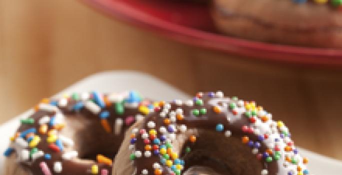 Fotografia em tons de marrom, branco e vermelho de uma bancada de madeira, sobre ela um prato branco com um donuts. Ao fundo um prato vermelho com donuts.