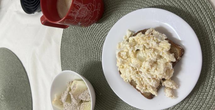 Fotografia de um prato raso médio branco com uma fatia de pão e ovos mexidos cremosos com farelo de aveia. Ao lado tem um recipiente pequeno de vidro e branco com melão e aveia e uma xícara vermelha grande.