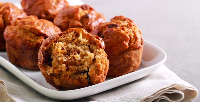 Fotografia de alguns muffins de maçã sobre um apoio retangular branco, o qual está apoiado em um paninho também branco. O pano está sobre uma bancada de cimento queimado claro.