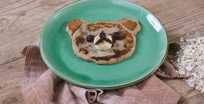 Foto da receita de Panqueca de Banana. Observa-se um pratinho verde e a panquequinha com cara de ursinho.