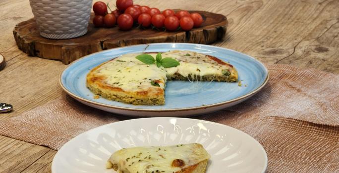 Fotografia em tons de marrom com um prato azul ao centro. Em cima do prato existe uma pizza com massa de Aveia coberta com queijo muçarela e orégano.