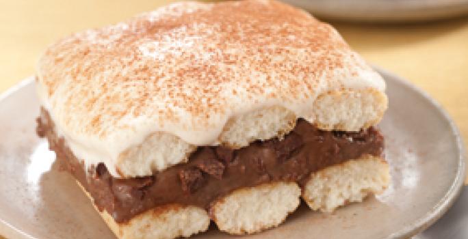 Fotografia em tons de bege em uma mesa de madeira com uma toalha amarela e um prato bege redondo raso ao centro com um pedaço de pavê de chocolate.