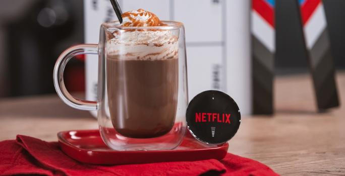 Foto em tons de vermelho e preto da receita de cappuccino netflix servida em um copo largo sobre uma base de porcelana vermelha e um pano vermelho em cima de uma mesa de madeira com uma cápsula de cappuccino netflix ao lado