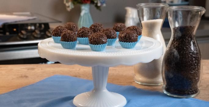 Fotografia de dez forminhas azuis com brigadeiros passados no granulado e apoiados em um enfeite para bolo branco e alto. Ao fundo tem um fogão, um vaso azul com flores, e ao lado do brigadeiro tem duas jarras decorativas de vidro com açúcar e cacau.