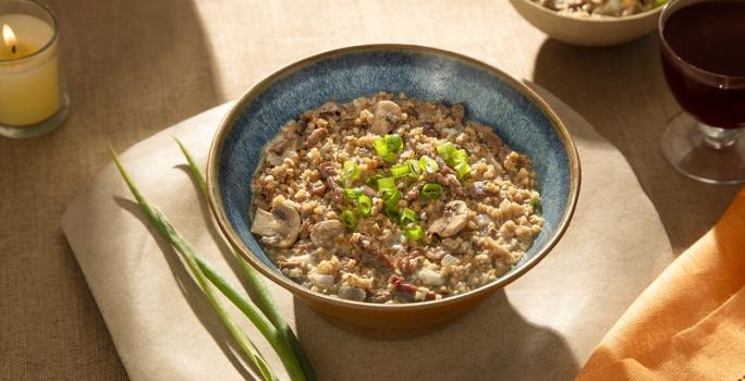 Foto em tons de marrom da receita de risoto de filé mignon e gorgonzola servida em um bowl de cerâmica azul sore um pano bege com ramos de alho poró ao lado