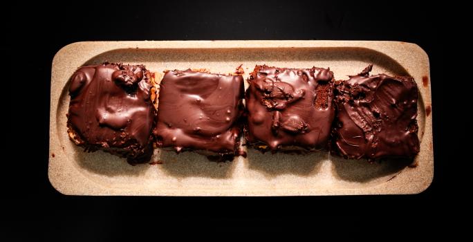 Fotografia vista de cima de quatro porções unidades de pão de mel com ganache de pitanga e cobertura de chocolate amargo. A sobremesa está em um apoio de madeira decorativo retangular, o qual está sobre uma mesa preta.