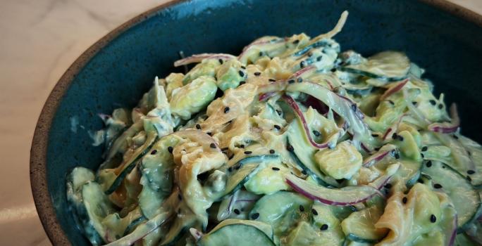 Fotografia em tons de marrom escuro com um bowl escuro ao centro. Dentro do bowl existe uma receita de salada de pepino feita com cream cheese, salmão, cebola e gergelim preto