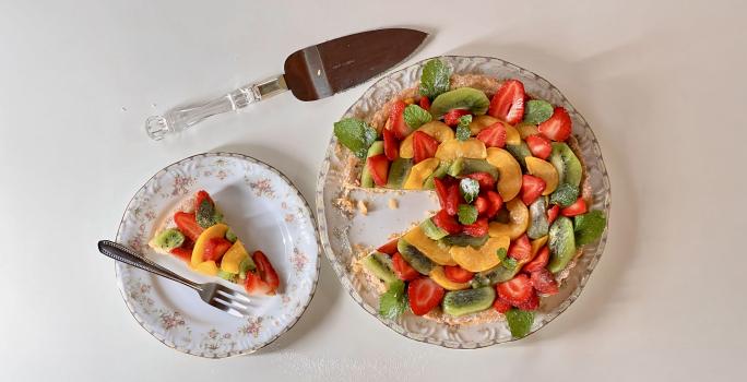 Foto vista de cima da receita de torta de frutas com creme de amêndoa, servida em um prato redondo ao lado de uma fatia cortada. A torta é decorada com frutas diversas (kiwi, manga e morango)