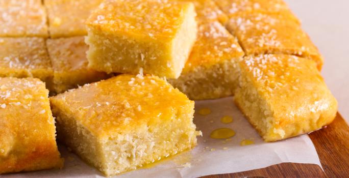 Fotografia em tons de amarelo com uma tábua ao centro. Em cima da tábua existe vários pedaços de bolo de cor amarela ao centro. Os pedaços de bolo estão cobertos com uma calda e coco ralado