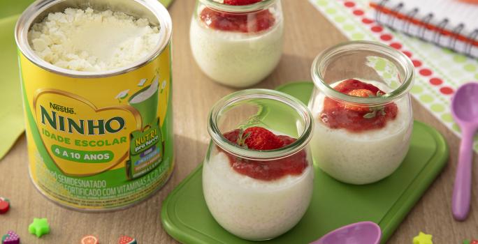 Fotografia de três recipientes de vidro pequenos com pudim de tapioca de ninho um creme de morango por cima. Dois desses recipientes estão sobre um apoio verde retangular, ao lado de uma colher roxa pequena e de uma lata de ninho escolar.