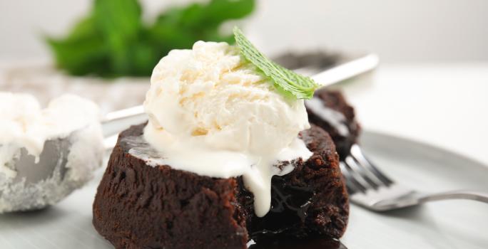 Fotografia de um petit gateau com uma bola de sorvete por cima e uma folha de hortelã. A sobremesa está em um prato raso e grande, com um garfo segurando um pedaço do bolinho e uma colher de sorvete meio cheia.