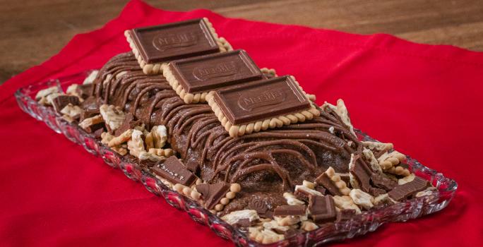 Fotografia de uma torta gelada de chocolate com chocolate derretido e alguns chocobiscuit por cima e ao lado.