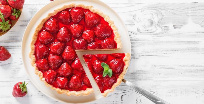 Fotografia vista de cima de um prato grande branco servindo como apoio para uma torta de morango zero lactose, com morangos por cima e uma calda brilhante. Tem um talher tirando um pedaço perfeito, e ao redor da torta tem vários morangos.