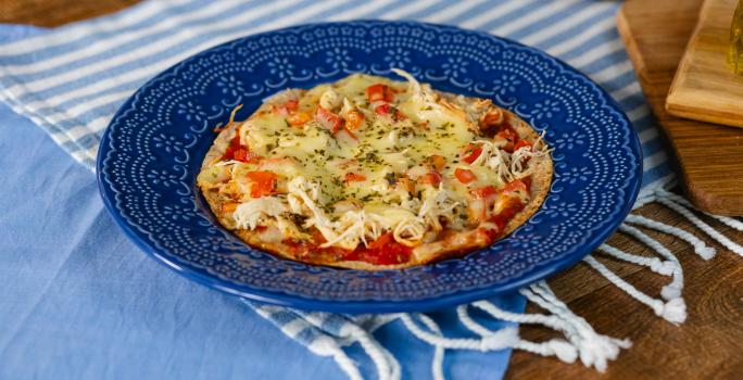 Fotografia em tons de azul com um prato azul ao centro. Em cima do prato existe uma receita de pizza de frigideira feita com frango desfiado, molho de tomate, queijo muçarela e orégano.