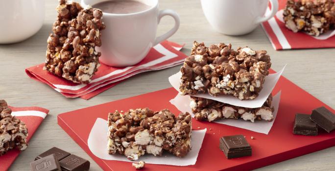 Fotografia em tons de vermelho em uma bancada de madeira clara com barras de pipoca com chocolate espalhadas pela mesa e em cima de uma tábua vermelha. Ao fundo, duas xícaras de chocolate quente com uma barra de pipoca com chocolate apoiada em uma delas.