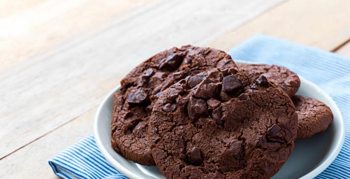 Fotografia de quatro cookies de chocolate com gotas de chocolate dentro de um recipiente de vidro branco um pouco fundo. O recipiente está por cima de um paninho azul com listras brancas sobre uma mesa de madeira.