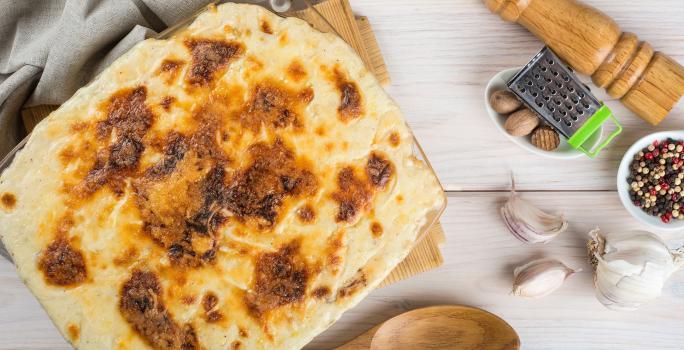 Fotografia de uma travessa de vidro quadrada de bacalhau gratinado com batata sobre uma tábua de madeira e um pano de tom cinza. No fundo tem um moedor de pimenta de madeira.