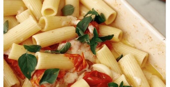 Fotografia de uma assadeira de cor clara com macarrão penne, tomates cereja assados, queijo feta derretido e folhas de manjericão.