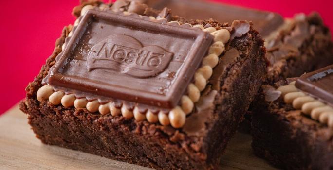 Fotografia três unidades de brownies com biscoito choco biscuit, eles estão apoiados um no outro e sobre uma tábua de madeira. O fundo da foto é vermelho.